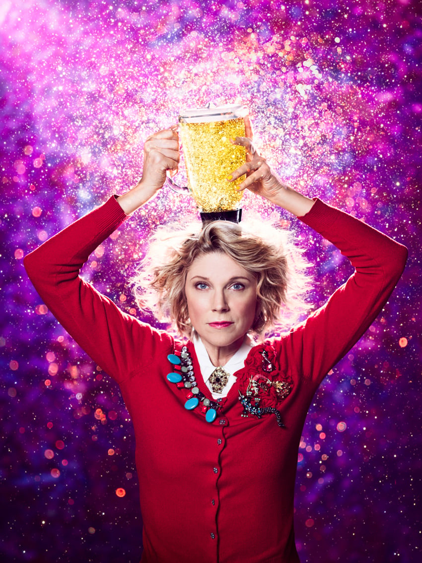 Cade Martin's photo for the theatre company Woolly Mammoth. The photo features a woman with short blonde wavy hair and a neutral expression wearing a red cardigan with a jumble of interesting brooches around the collar. She holds a blender pitcher that is seemingly filled with gold glitter on top of her head and stands against a glittery purple background.